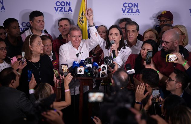 Mulher discursa em microfone, homem com braço erguido ao lado, cercados por pessoas e microfones de imprensa. Banner 'CON VZLA'.