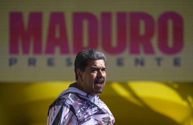 Nicolás Maduro discursa, com a boca aberta, à frente de um painel amarelo com "MADURO" em rosa e "PRESIDENTE" abaixo.