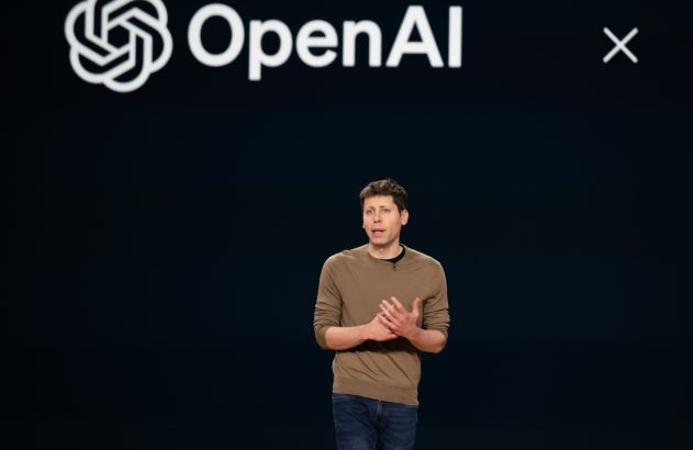 Palestrante gesticula em palco escuro com logo OpenAI no fundo.