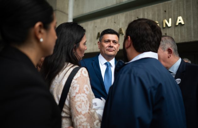 Homem de terno azul, foco central, entre pessoas. Ao fundo, prédio com letras "GNA".