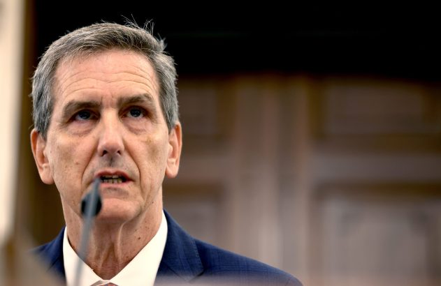 Close-up de homem com cabelo grisalho e terno falando ao microfone, olhando para cima.