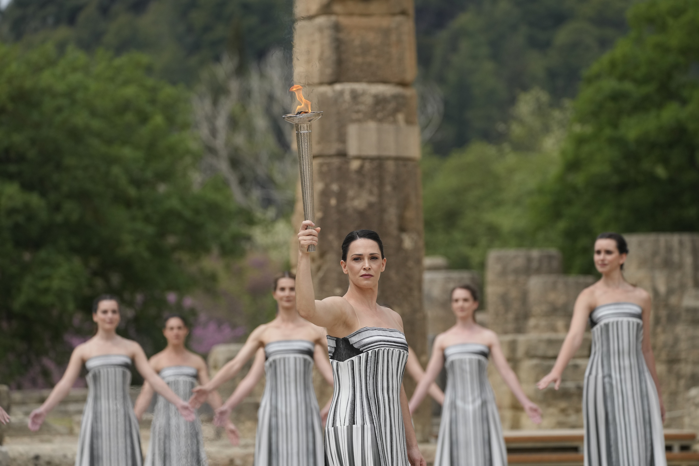 Mulher em vestido listrado cinza ergue tocha olímpica flamejante. Outras em similar vestuário em ruínas históricas.