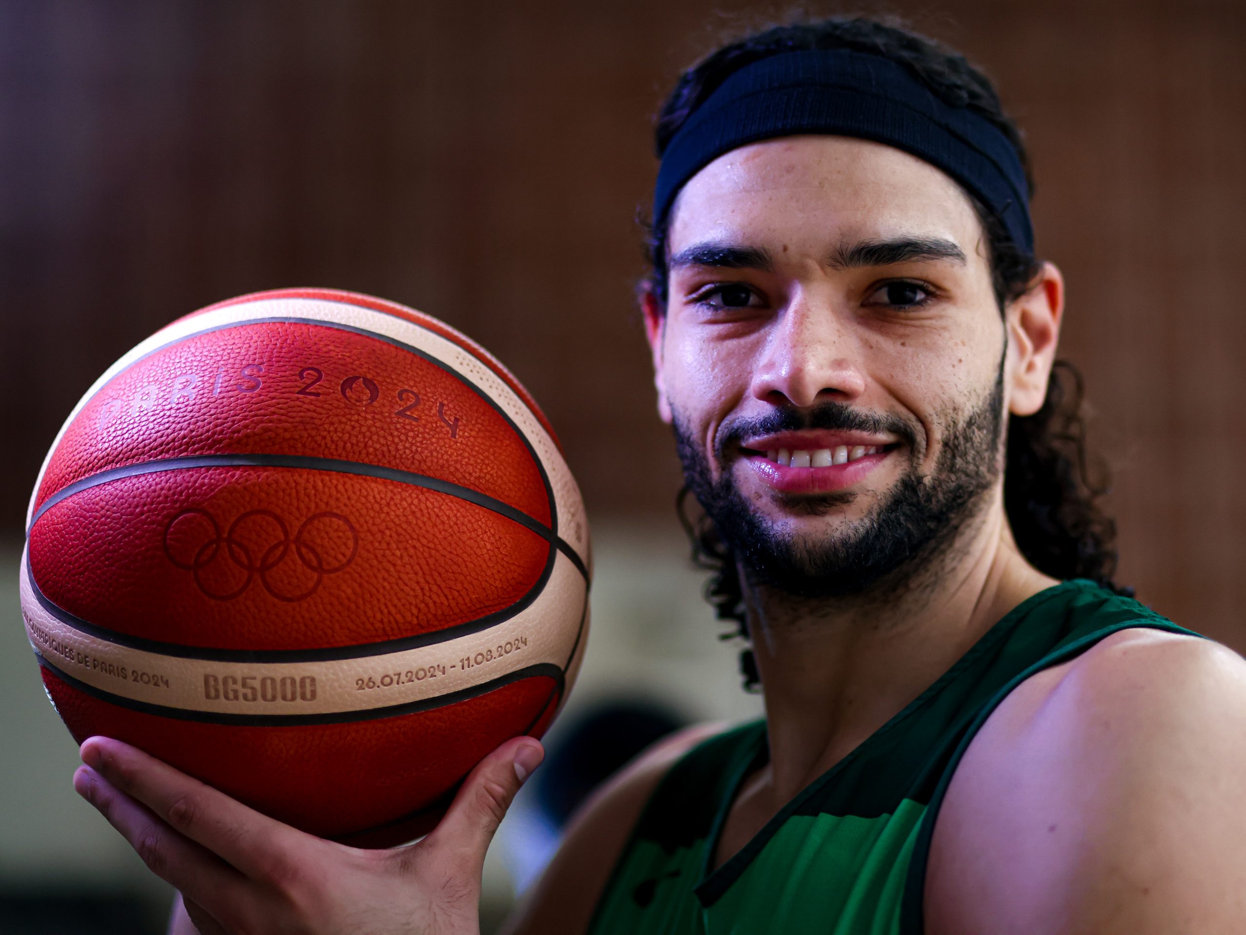 Homem sorrindo, segurando bola de basquete com logo "Paris 2024" e anéis olímpicos.