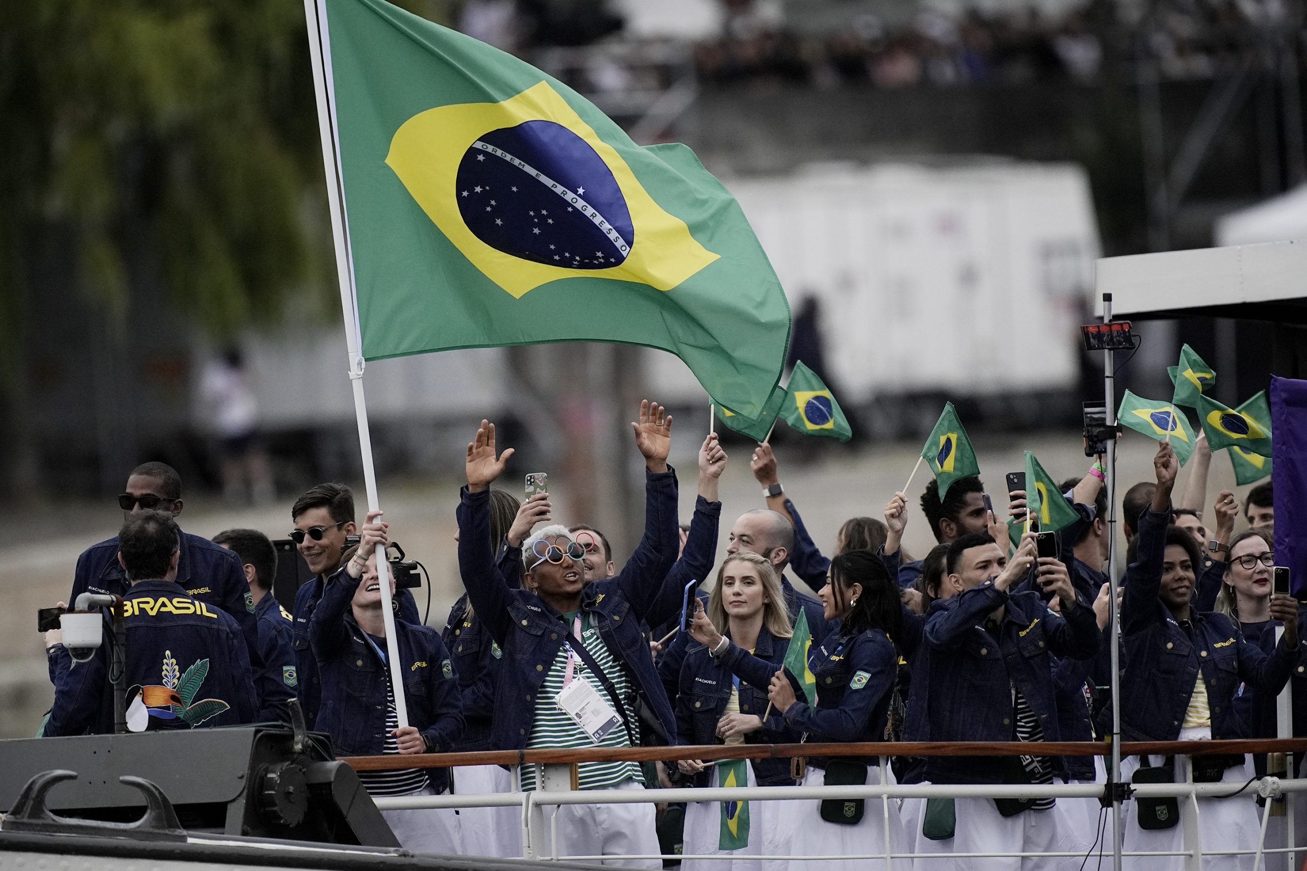 Delegação brasileira em barco, agitando bandeiras do Brasil e celebrando com as mãos para o alto.
