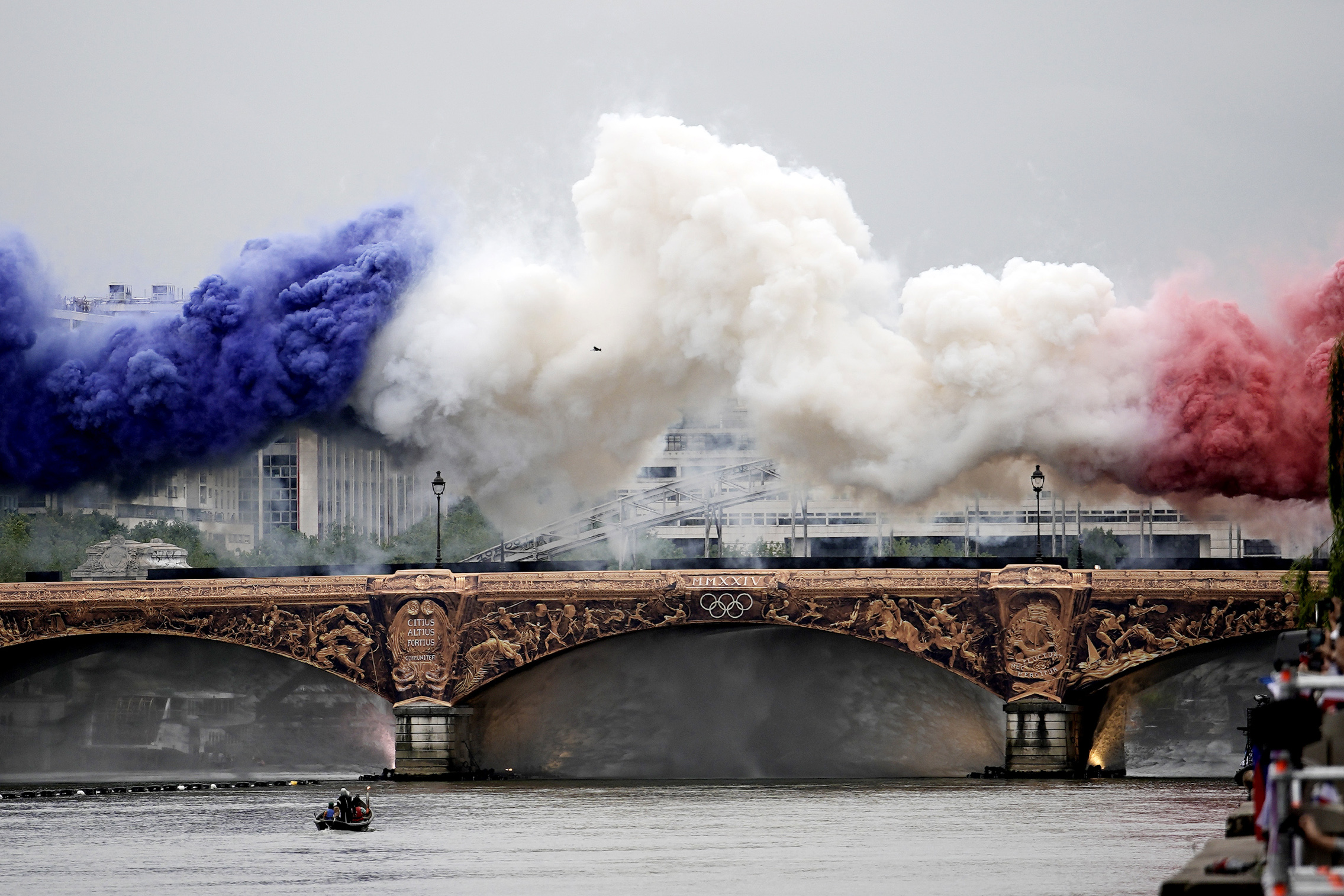 Fumaça azul, branca e vermelha sobre a Ponte Alexandre III no rio Sena, com anéis olímpicos.