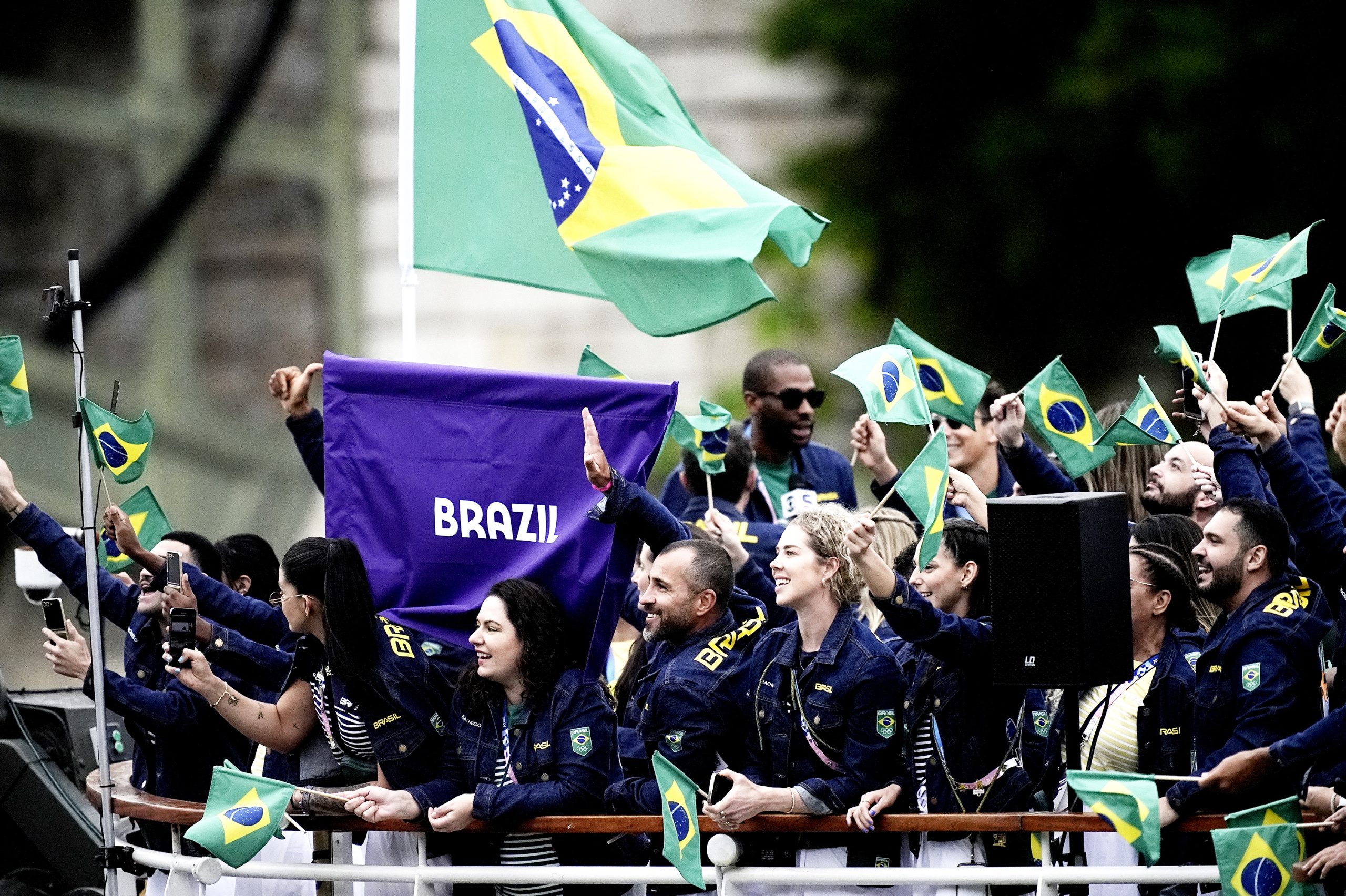 Pessoas em um desfile agitam bandeiras do Brasil. Um banner roxo com "BRAZIL" se destaca.