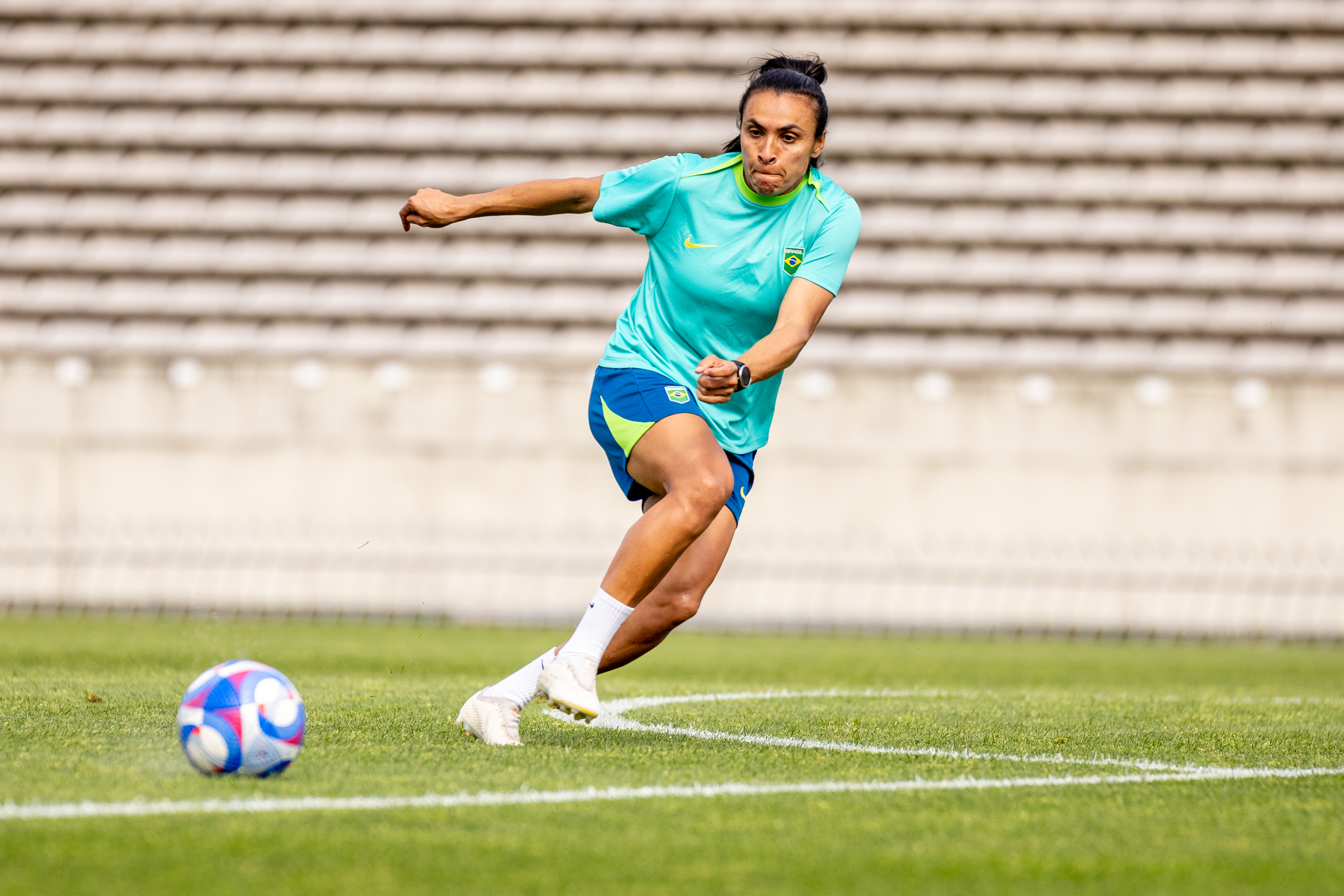 Jogadora brasileira de futebol, com uniforme azul e turquesa, correndo no campo com uma bola ao lado.