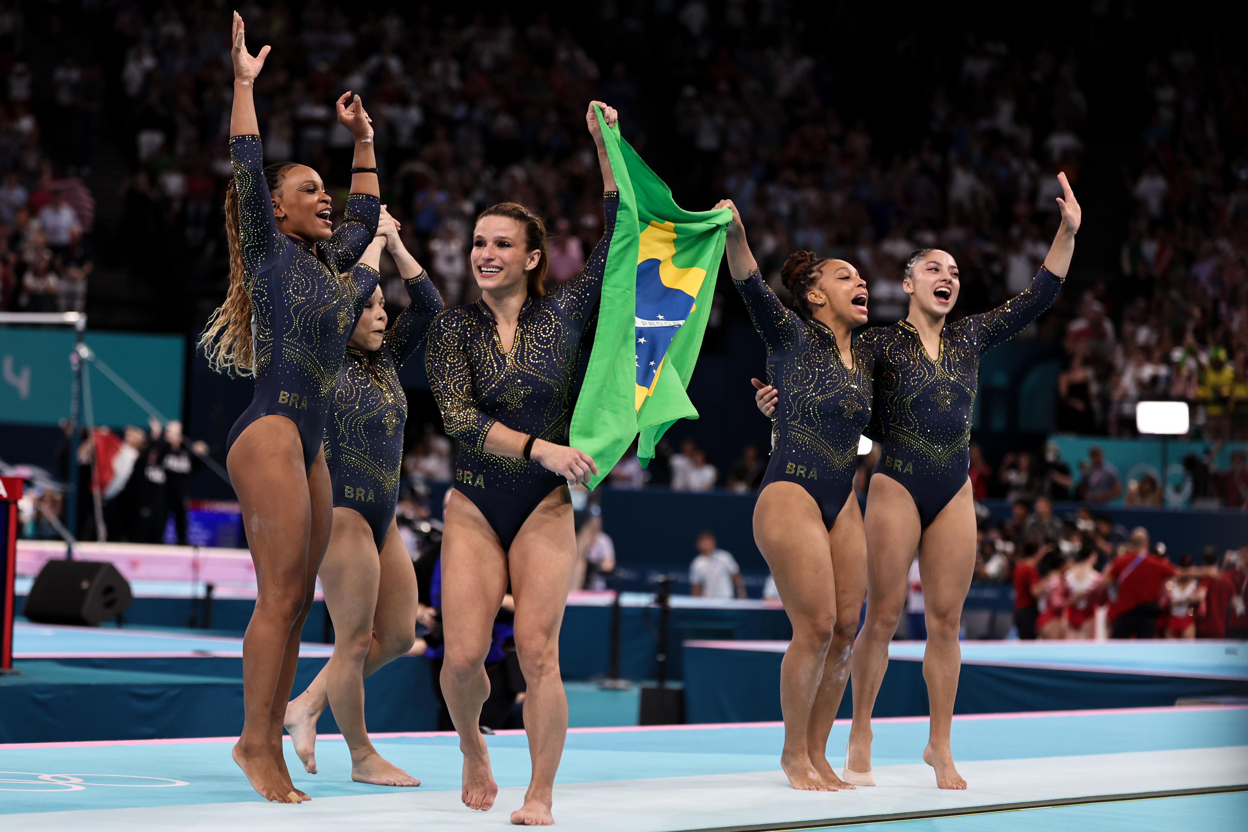 Ginastas brasileiras comemoram, uma segurando a bandeira do Brasil.
