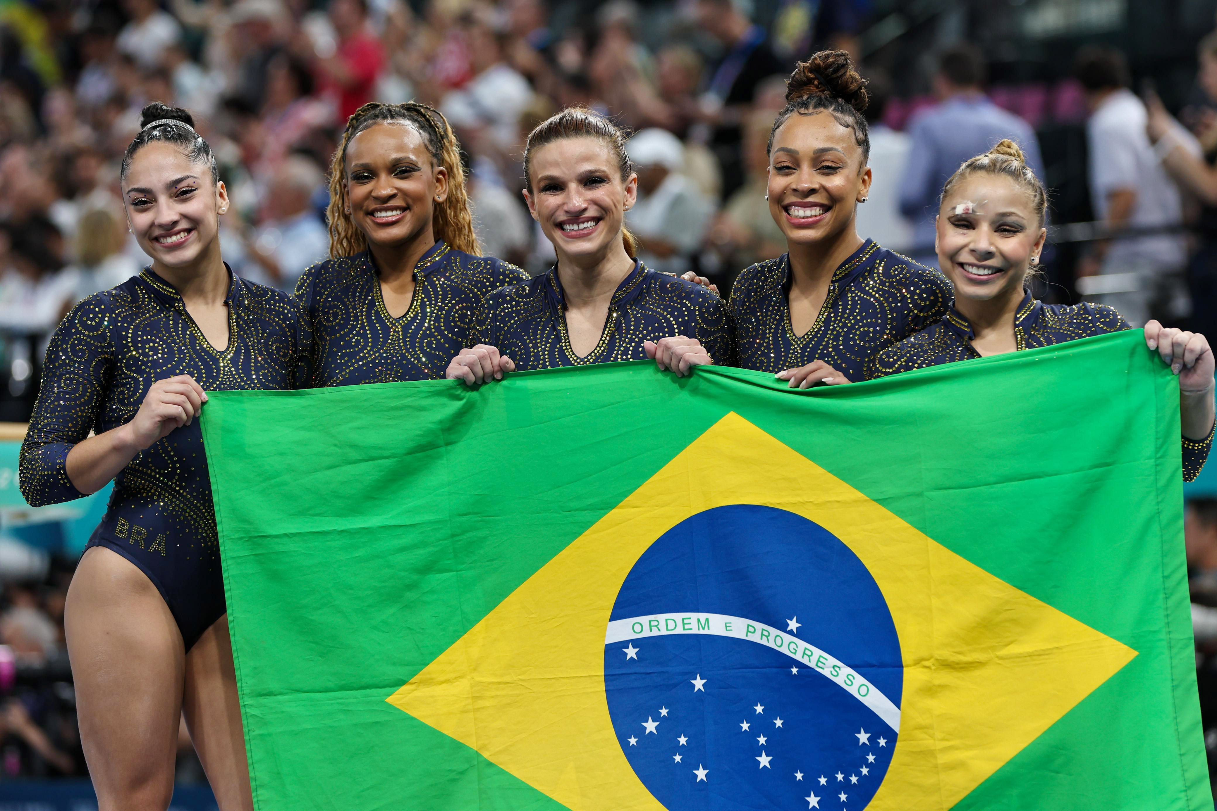 Equipe brasileira de ginástica sorri ao exibir a bandeira do Brasil.
