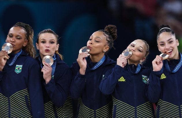 Cinco ginastas brasileiras em uniformes azul marinho seguram medalhas de bronze, algumas beijando-as, com sorrisos de celebração.