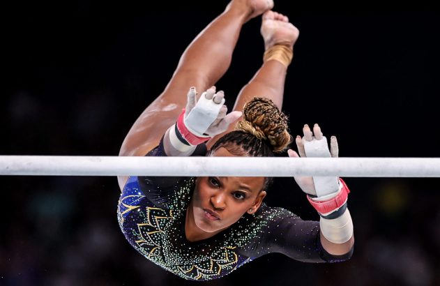 Ginasta em barra assimétrica, de cabeça para baixo, com collant brilhante e protetores de mão, olhando para frente.