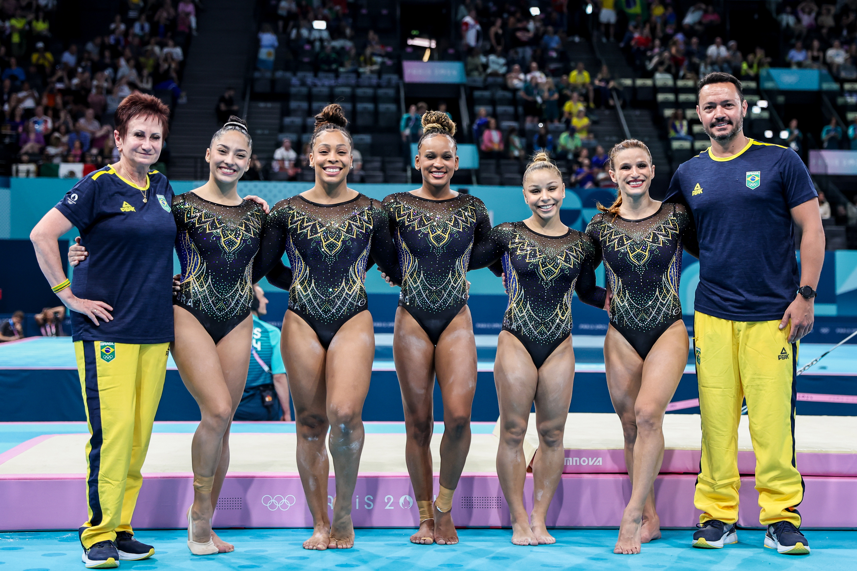 Equipe de ginástica do Brasil: cinco ginastas e dois treinadores sorriem em foto de grupo no ginásio.