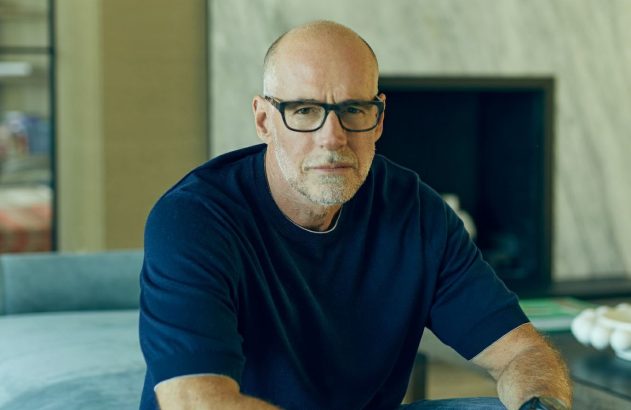 Homem careca de barba grisalha e óculos, com camisa azul, sentado, olhando fixamente para a câmera.
