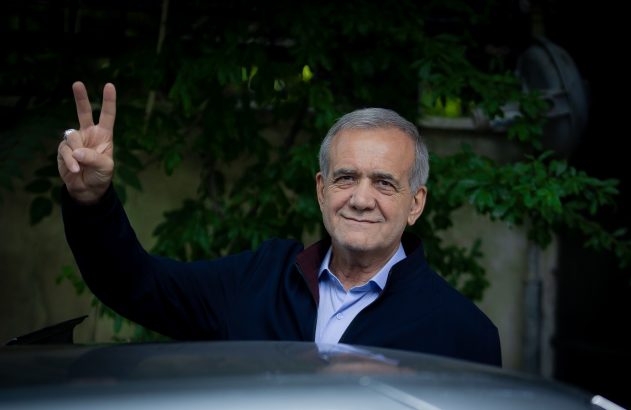 Homem sorridente levanta a mão em sinal de vitória, com folhagem verde ao fundo e o topo de um carro.
