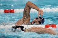 Nadador respirando em nado crawl, anéis olímpicos tatuados no braço, com água espirrando em piscina.