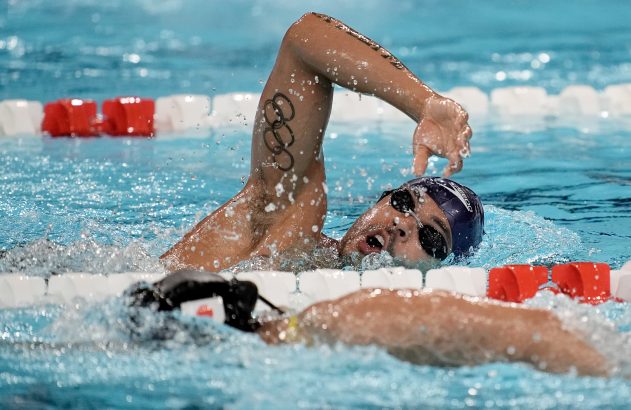 Nadador respirando em nado crawl, anéis olímpicos tatuados no braço, com água espirrando em piscina.
