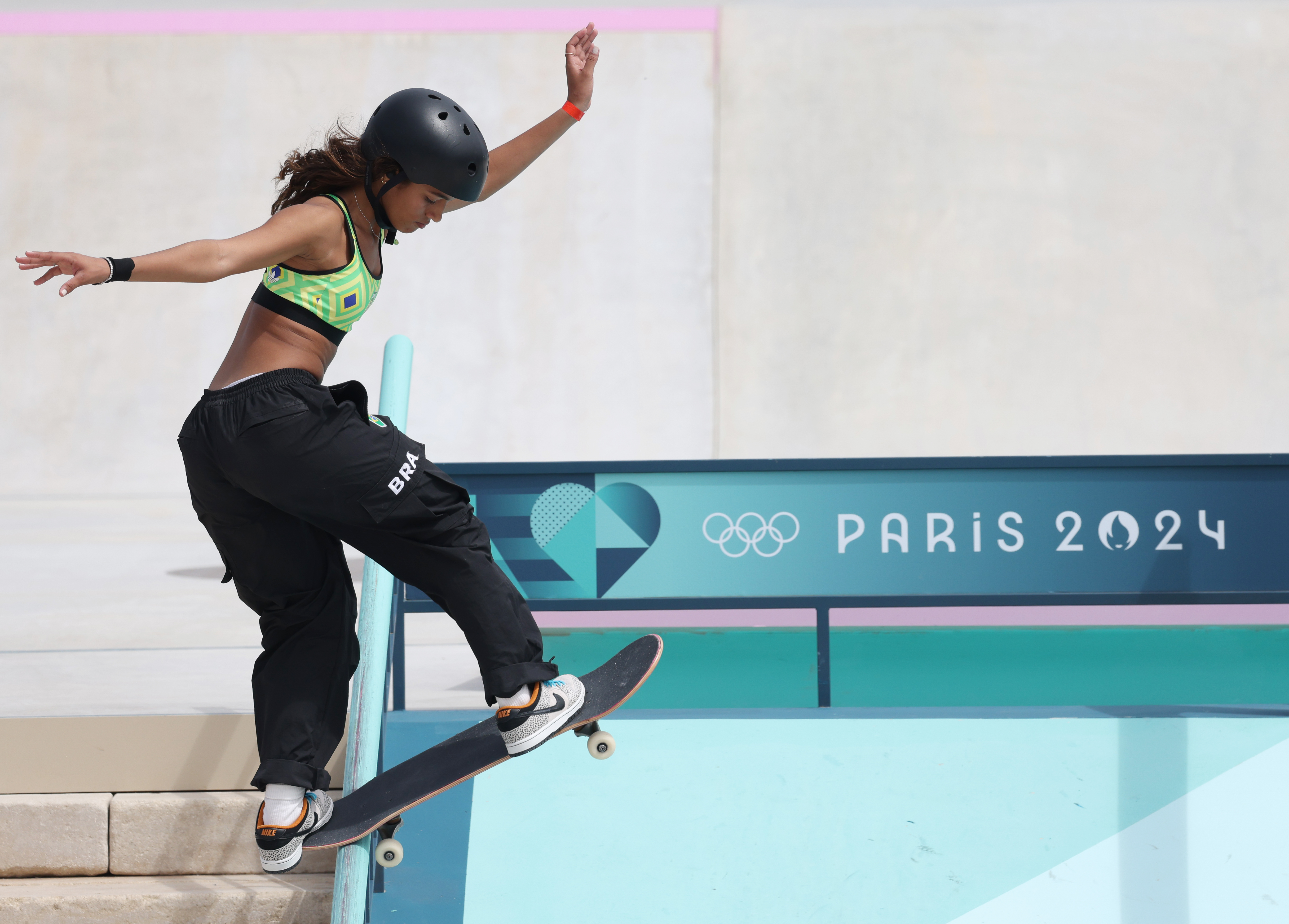 Mulher skatista com capacete e top verde realizando manobra em pista do Paris 2024.