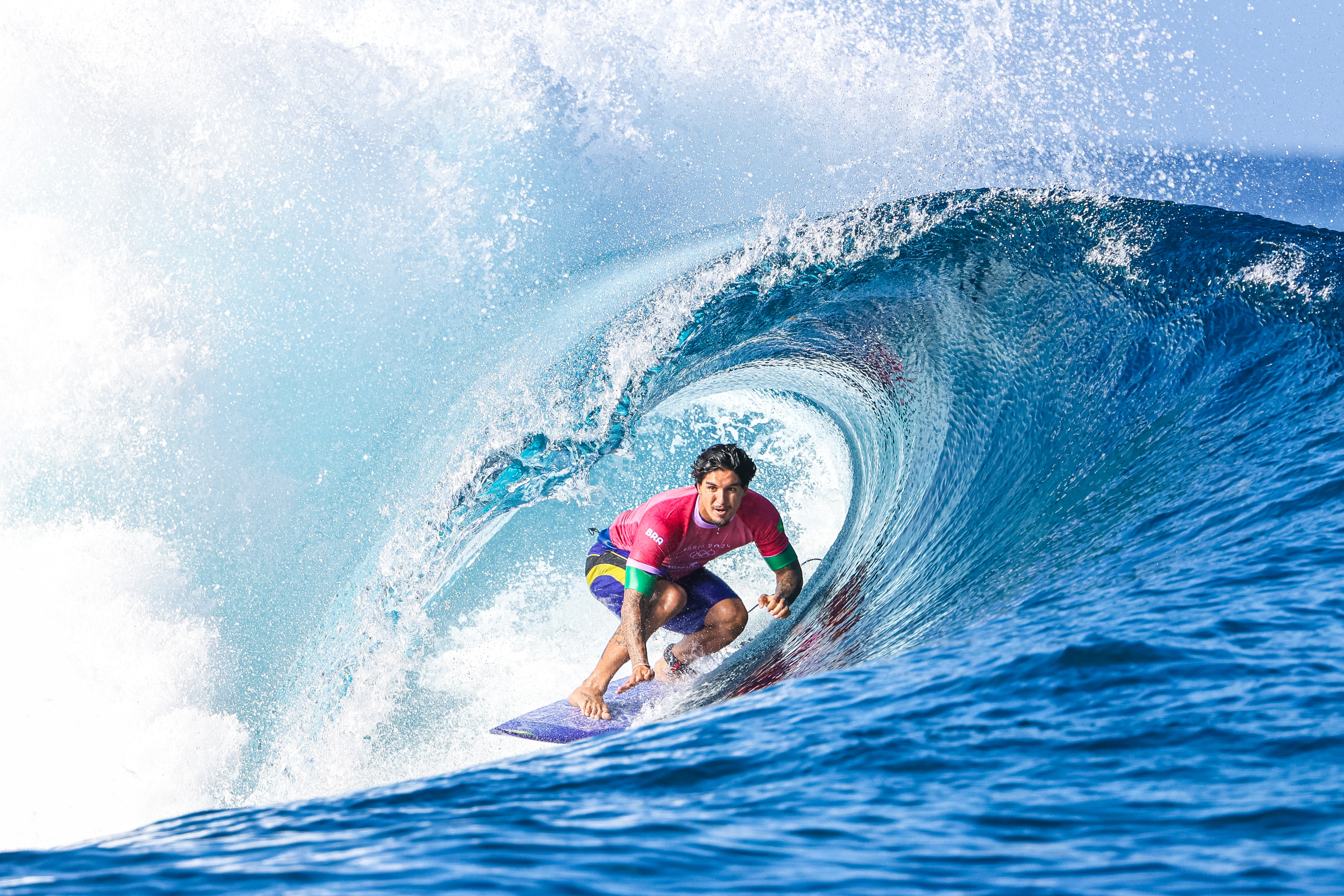 Surfista de camisa rosa surfando dentro de um tubo de onda azul, com espuma e spray.