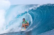 Surfista com capacete e blusa verde, surfando uma onda tubular azul-turquesa, com água espirrando.