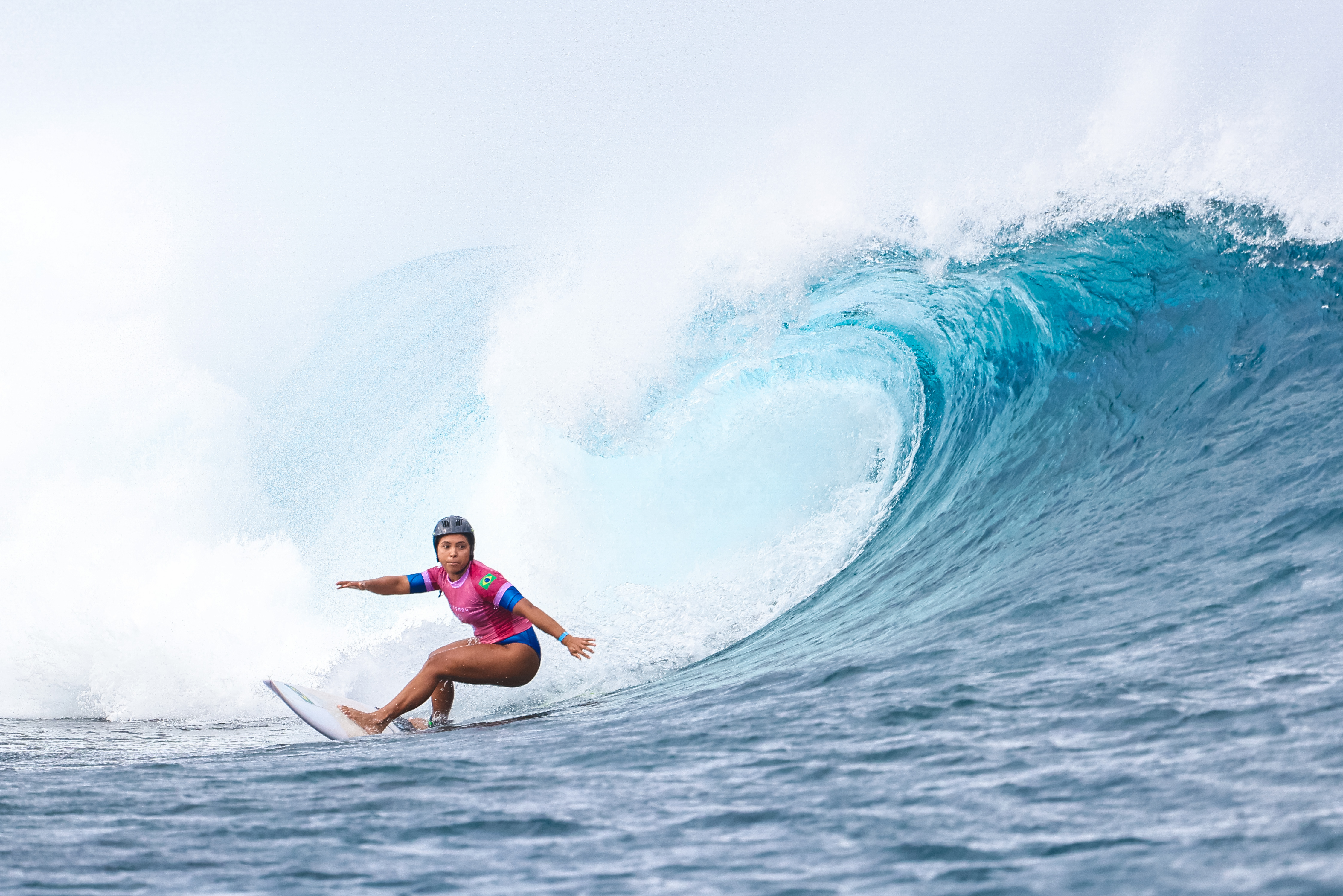 Mulher surfista de capacete desce grande onda tubular azul com espuma. Camisa com bandeira do Brasil.