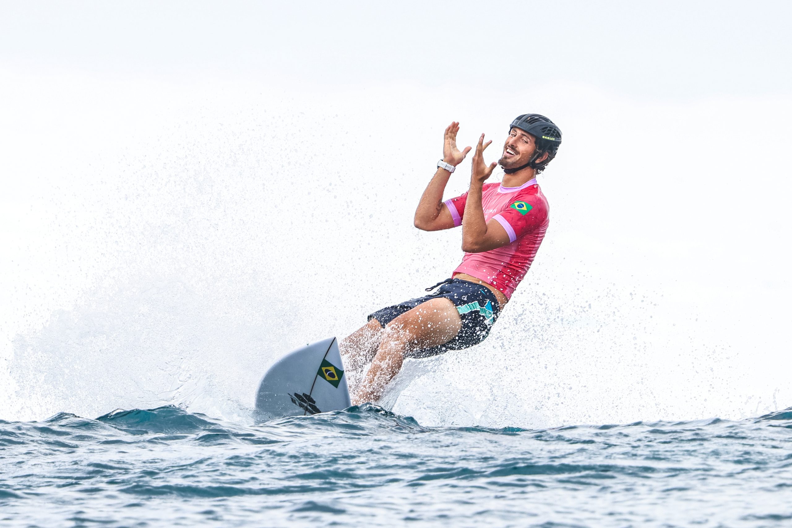Surfista de hidrofólio com capacete e camisa rosa, braços erguidos celebrando, água espirrando.