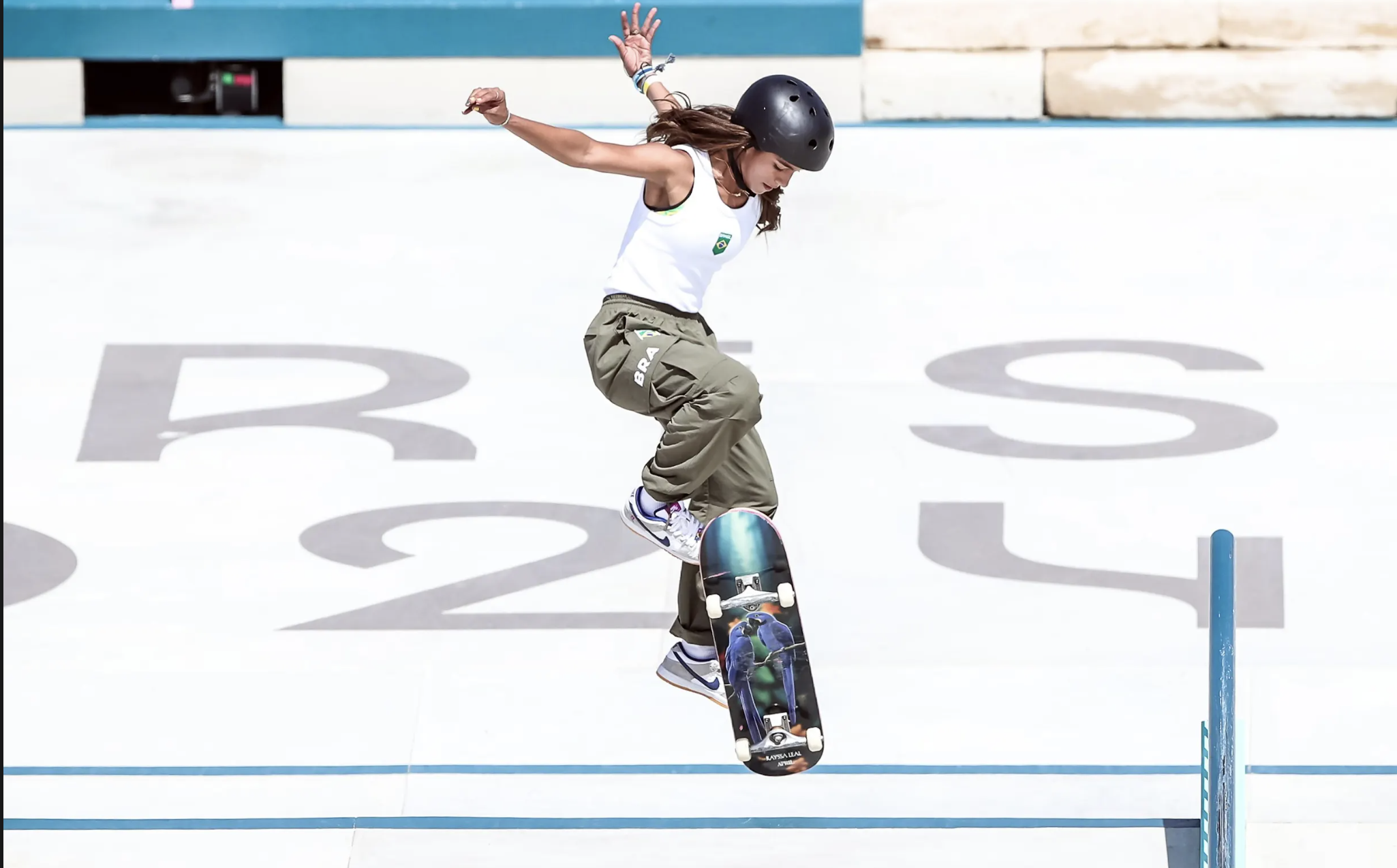 Skatista brasileira em manobra aérea. Skate com papagaios azuis. Fundo branco com 'BRAS'.