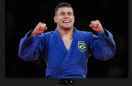 Judoísta brasileiro de quimono azul sorrindo e celebrando com punhos erguidos.