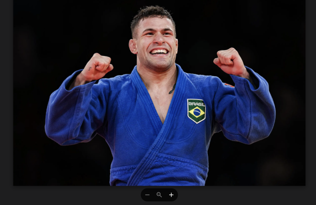 Judoísta brasileiro de quimono azul sorrindo e celebrando com punhos erguidos.