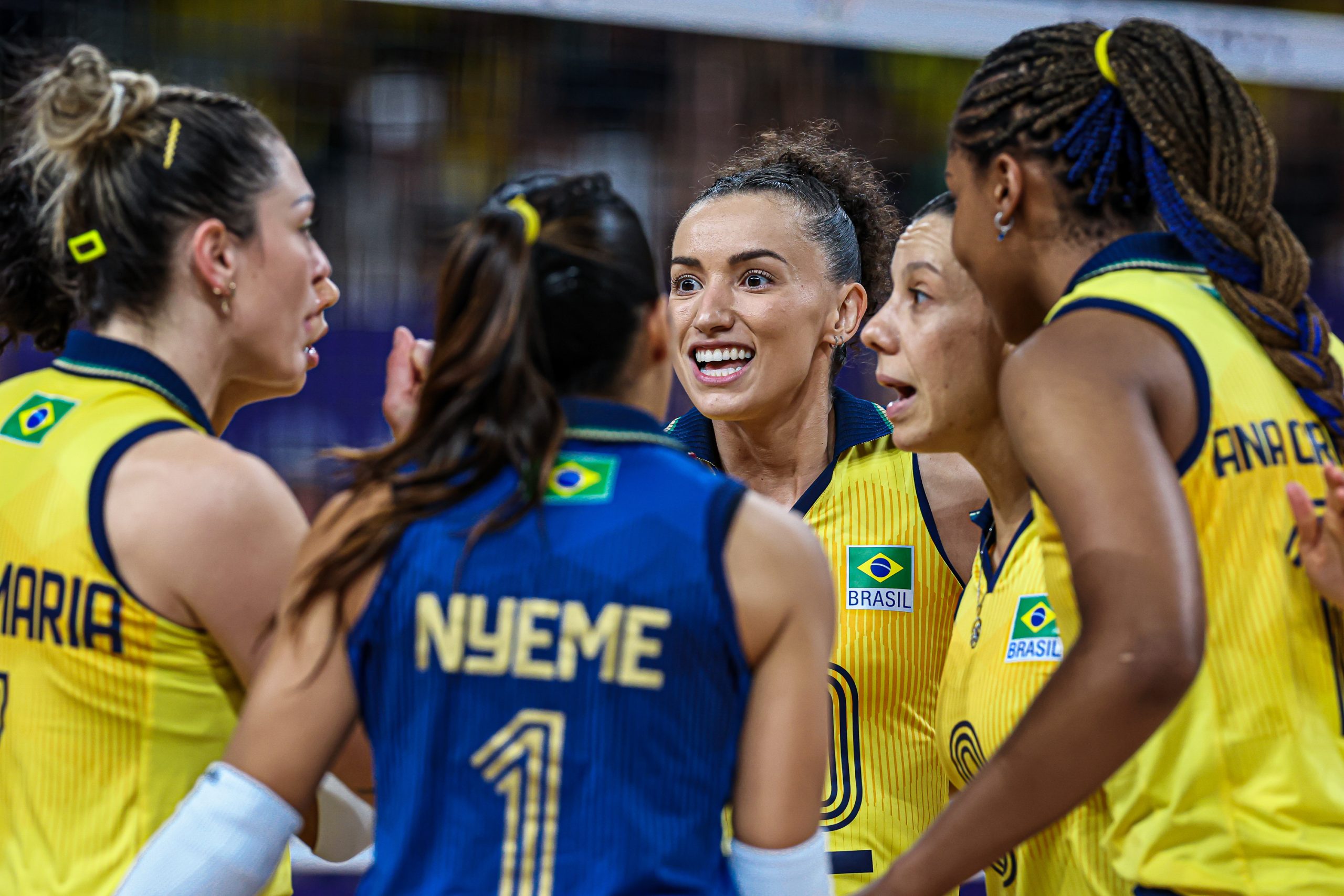 Jogadoras de vôlei do Brasil em roda, uma sorrindo no centro.