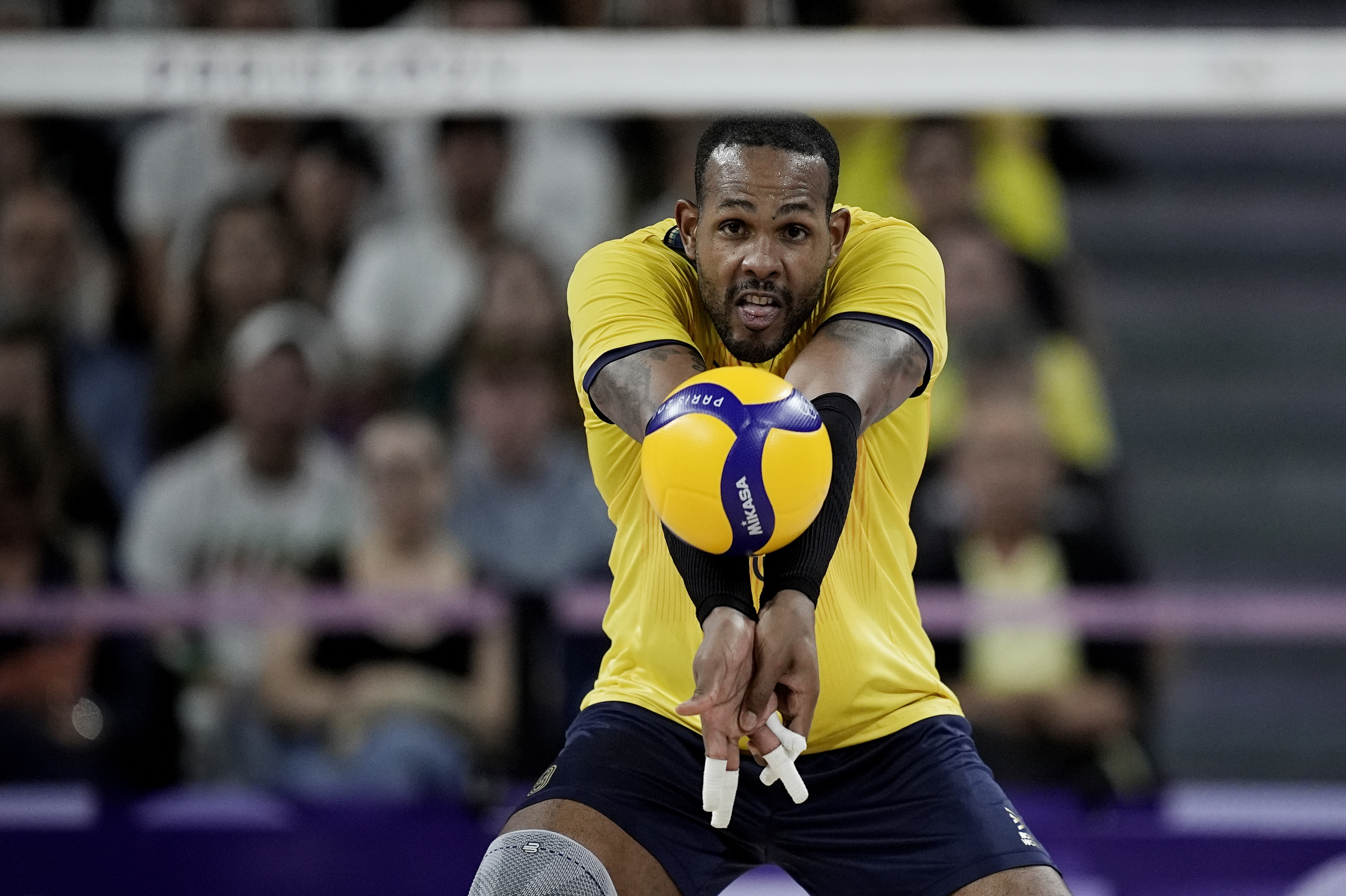 Jogador de vôlei, camisa amarela, realizando passe de manchete com bola amarela e azul. Quadra e torcida ao fundo.