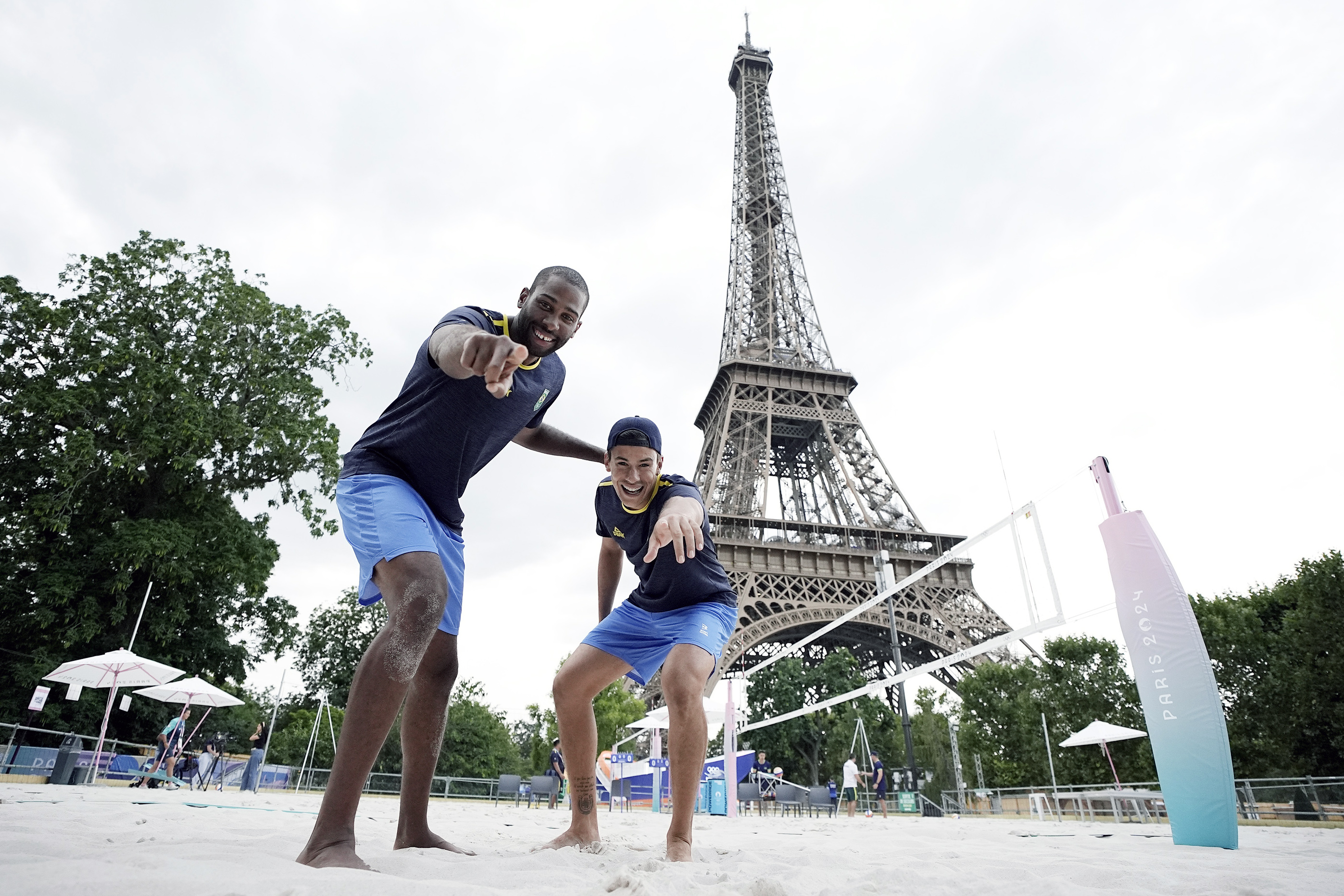 Dois atletas de vôlei de praia sorrindo e apontando na areia, com a Torre Eiffel ao fundo.