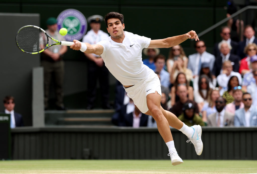 Tenista masculino de branco em salto, golpeando a bola com a raquete em uma quadra de grama de Wimbledon.