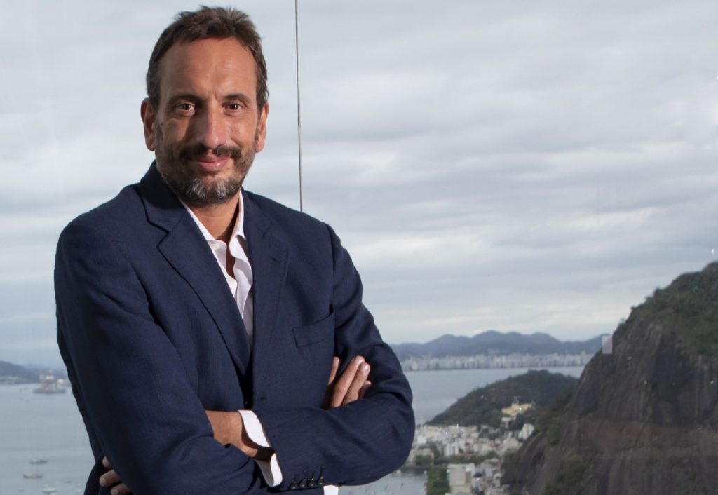 Homem sorrindo de terno com braços cruzados em frente a uma janela com vista do Rio de Janeiro.