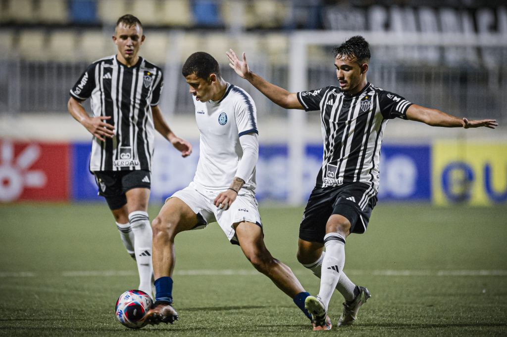 Três jogadores de futebol em campo. Jogador de camisa branca dribla a bola sob marcação de jogador com camisa listrada P&B.