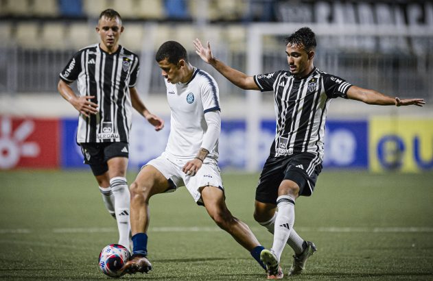 Três jogadores de futebol em campo. Jogador de camisa branca dribla a bola sob marcação de jogador com camisa listrada P&B.