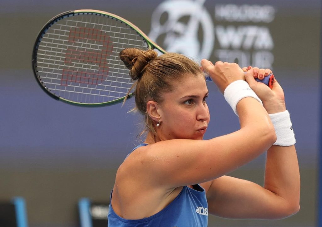 Tenista feminina concentrada, empunhando raquete, com camiseta azul e munhequeiras brancas. Fundo de quadra com logo WTA.