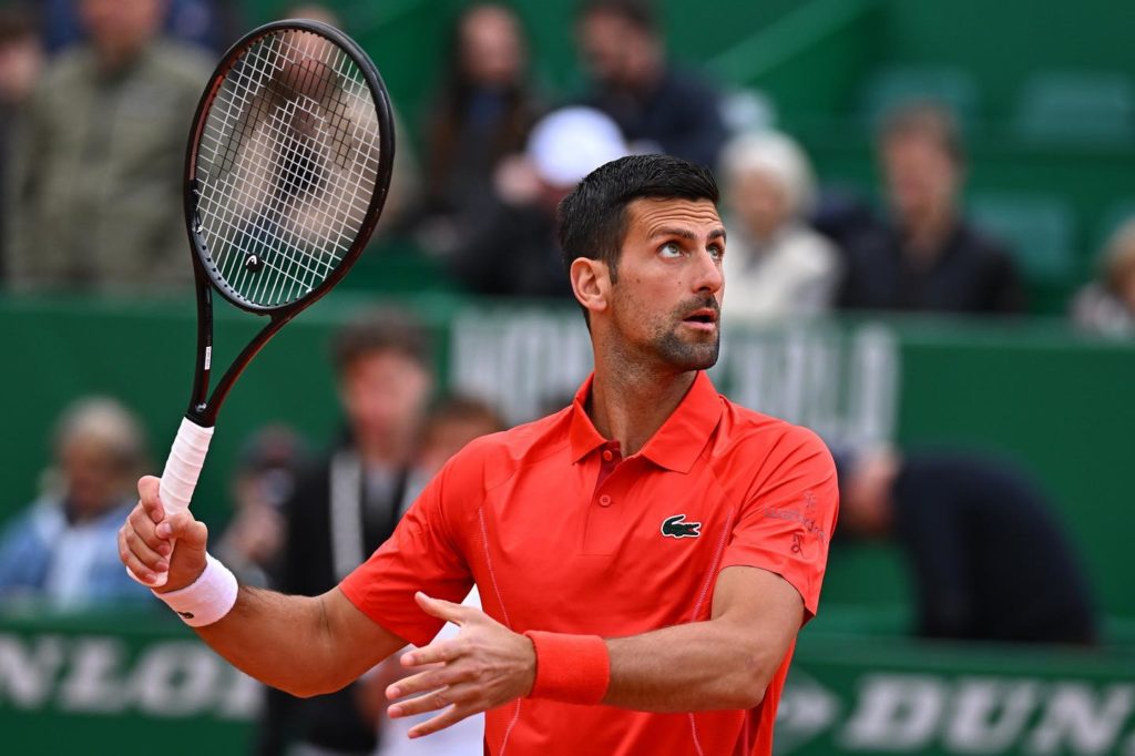Novak Djokovic em quadra de tênis, com raquete na mão e camisa vermelha, olhando para cima.