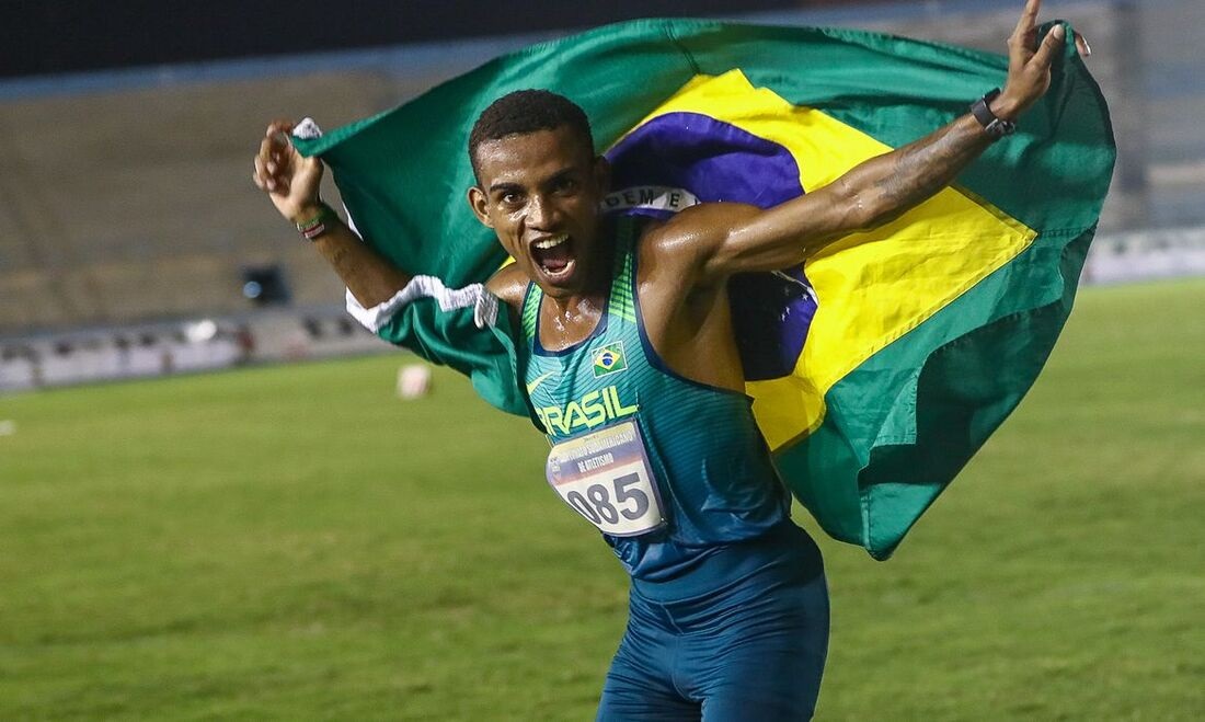 Atleta em pista comemora vitória, agitando a bandeira do Brasil como capa. Expressão de triunfo.