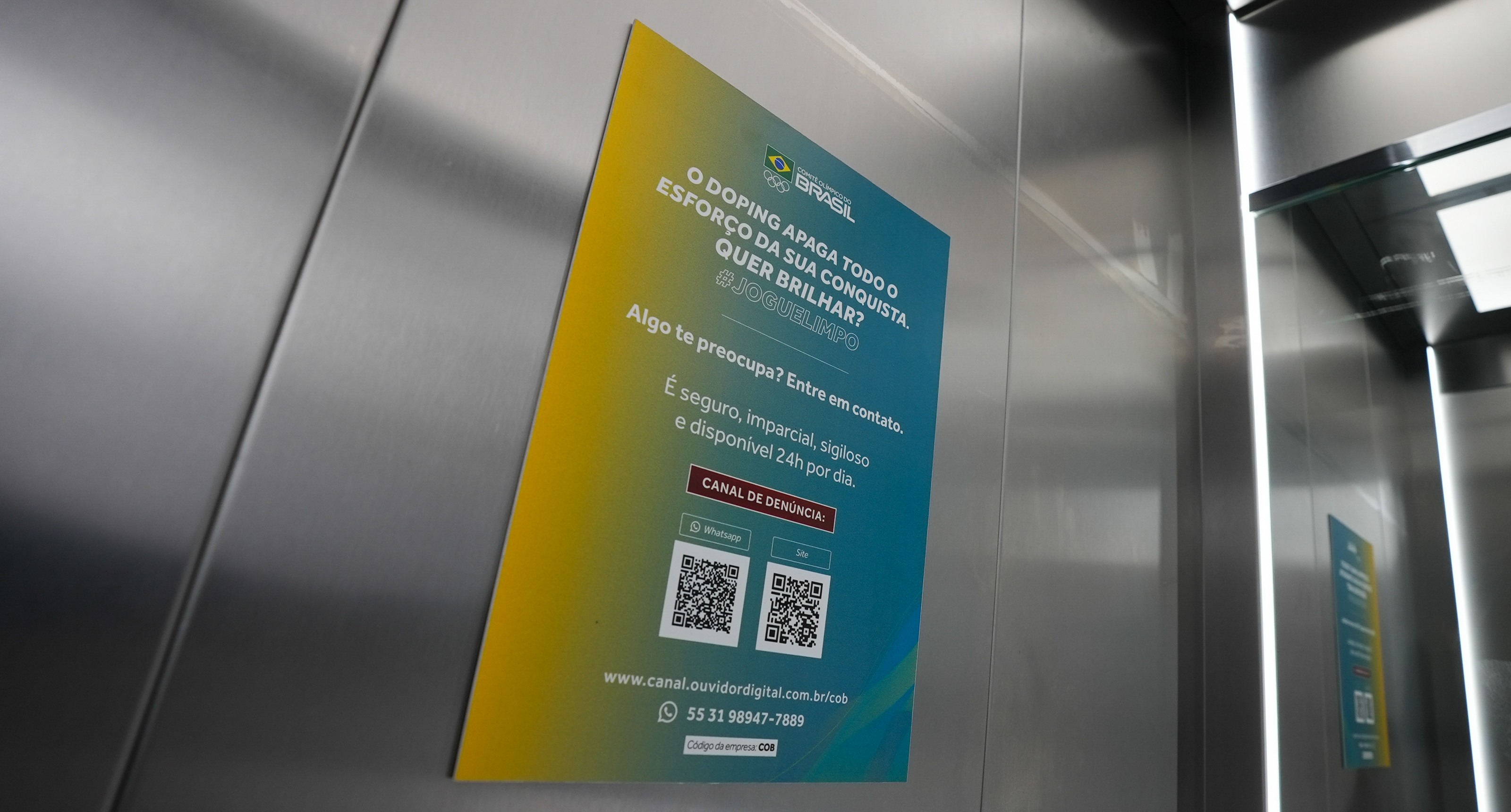 Cartaz em degradê amarelo e azul com texto e QR codes, afixado na parede metálica de um elevador.