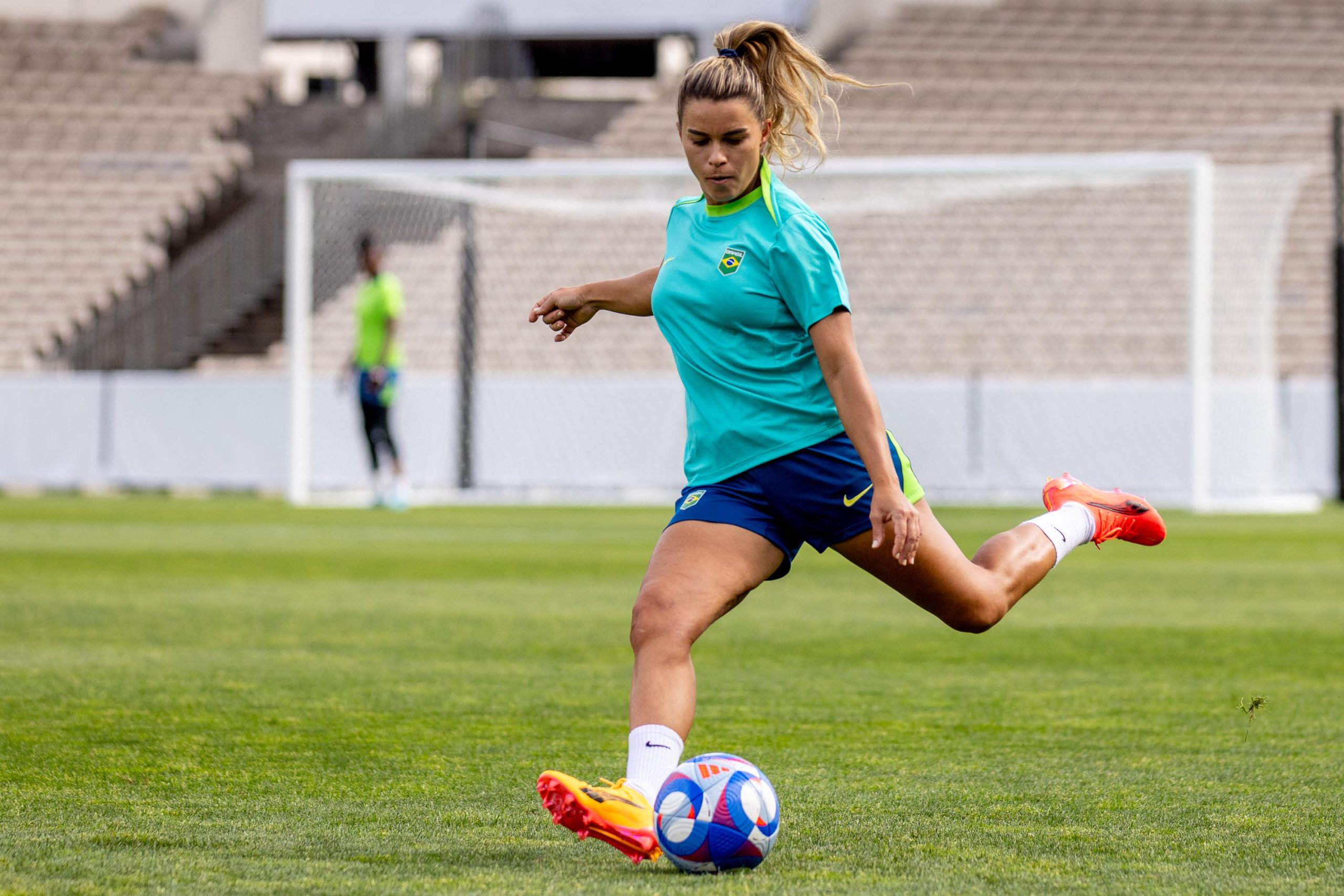 Jogadora chuta bola de futebol em campo de grama. Veste uniforme esportivo, estádio ao fundo.