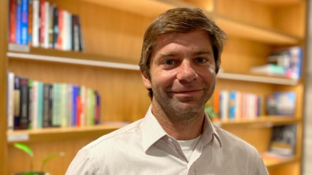 Um homem de camisa clara sorri para a câmera com estantes de livros em segundo plano.