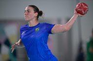 Atleta brasileira de handebol em uniforme azul, preparando-se para arremessar a bola vermelha.