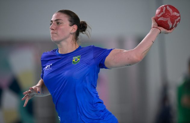 Atleta brasileira de handebol em uniforme azul, preparando-se para arremessar a bola vermelha.