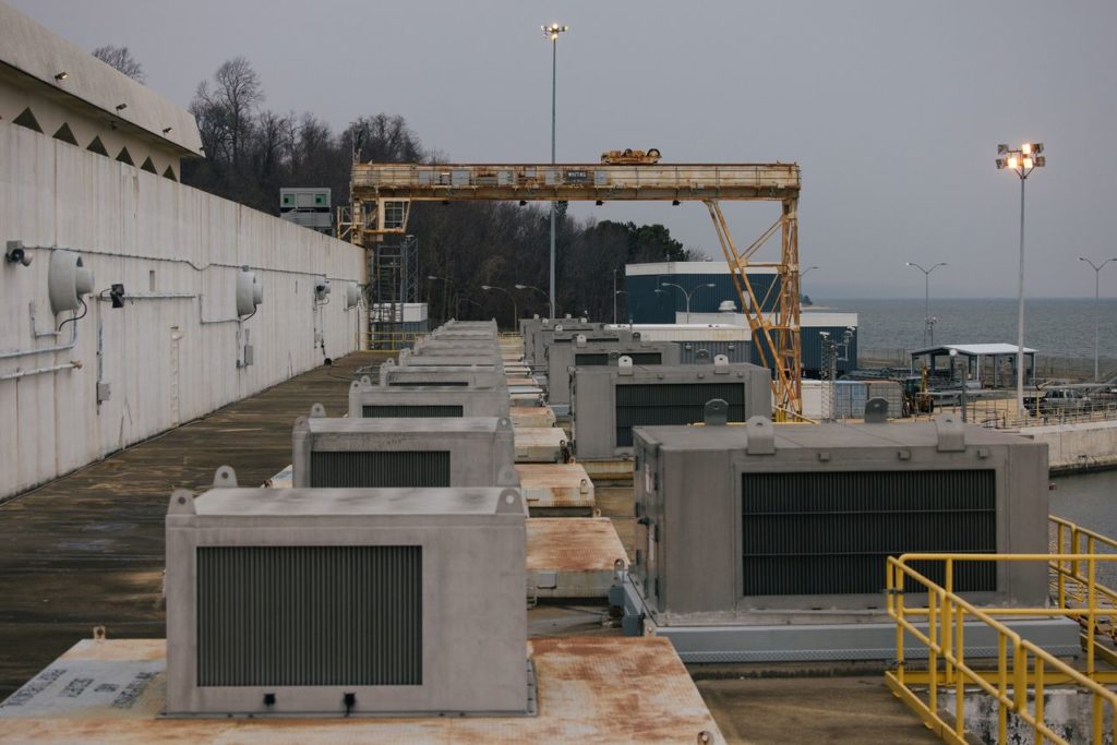 Longa fila de equipamentos industriais cinzas ao lado de muro branco. Guindaste e vasto corpo d'água ao fundo.