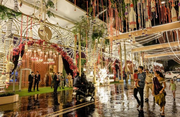 Pessoas em um salão grande e ricamente decorado com arranjos suspensos e chão refletivo, durante um evento.