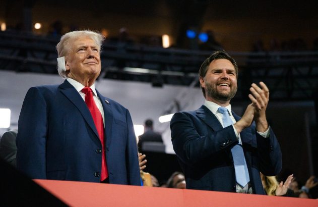 Donald Trump sorri e olha para cima ao lado de J.D. Vance, que sorri e aplaude em evento.