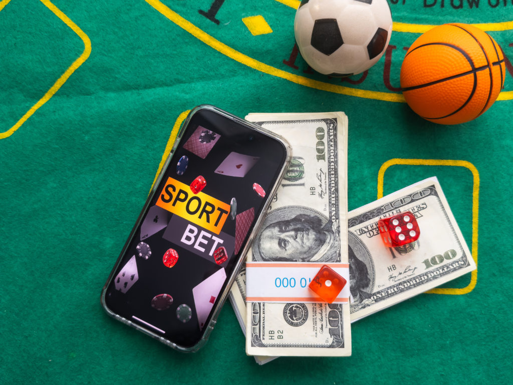 Smartphone com app "SPORT BET", dinheiro, dados e bolas de futebol/basquete em mesa de jogo verde.