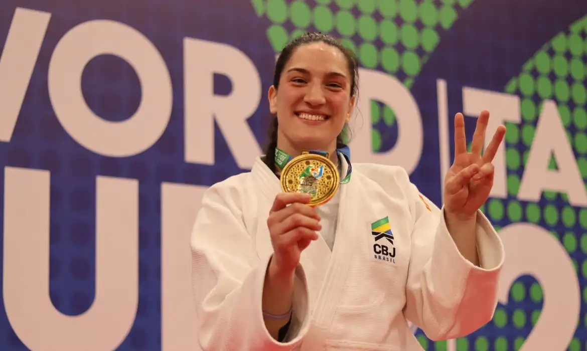 Judoca brasileira sorridente com medalha de ouro e três dedos levantados.