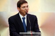 Homem de terno e óculos discursando ao microfone em púlpito.