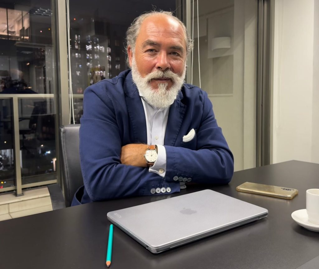 Homem com barba branca e blazer azul, sorrindo em seu escritório, com laptop e café na mesa.